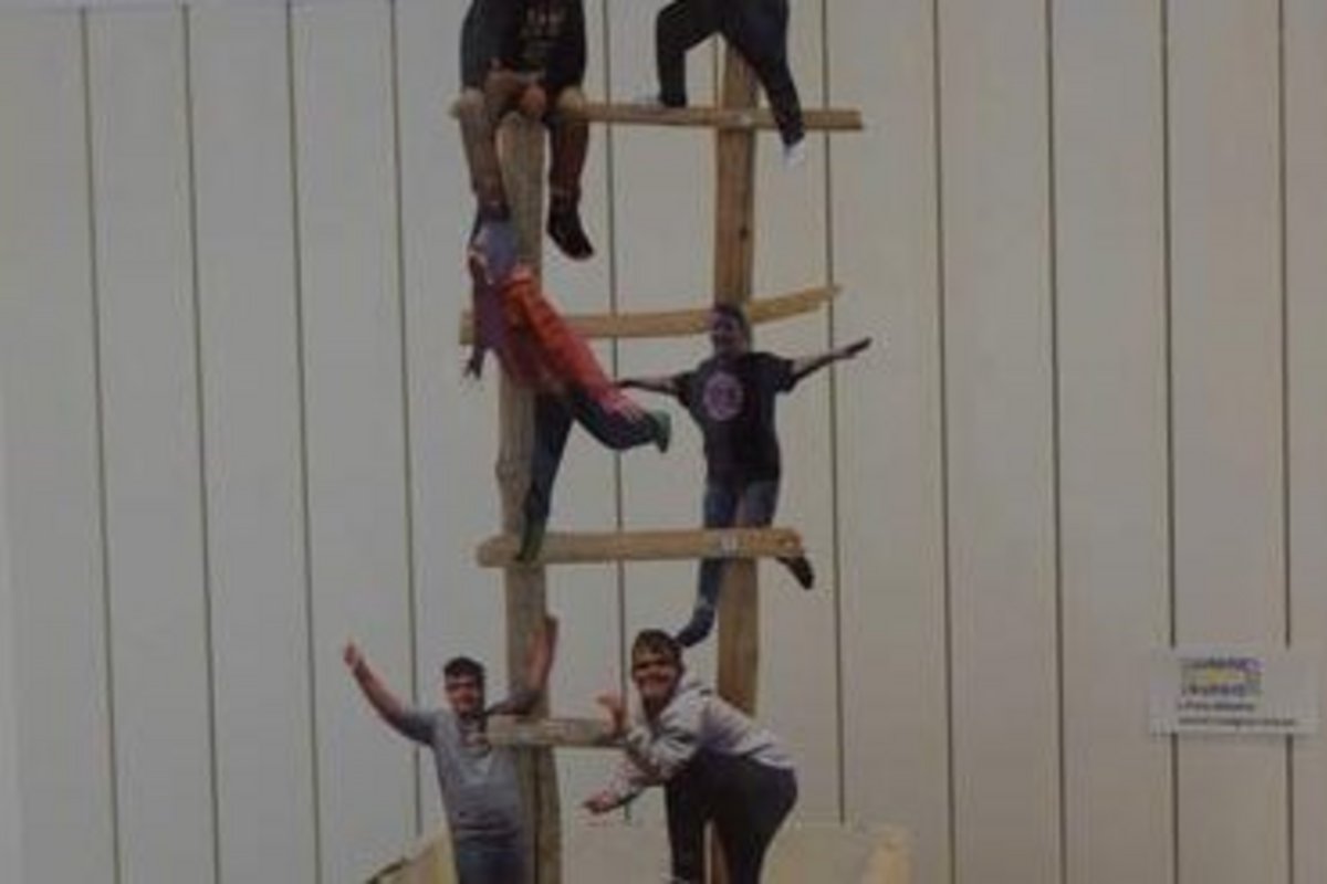 eine ca. 3 Meter hohe Holzleiter, die aus einer großen Holzschublade ragt und auf der mehrere Menschen sitzen