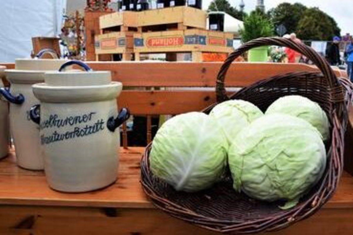 4 Kohlköpfe in einem geflochtenen Korb stehend auf einer Holzbank - neben dem Korb 2 Gefäße mit der Aufschrift "... Krautwerkstatt"